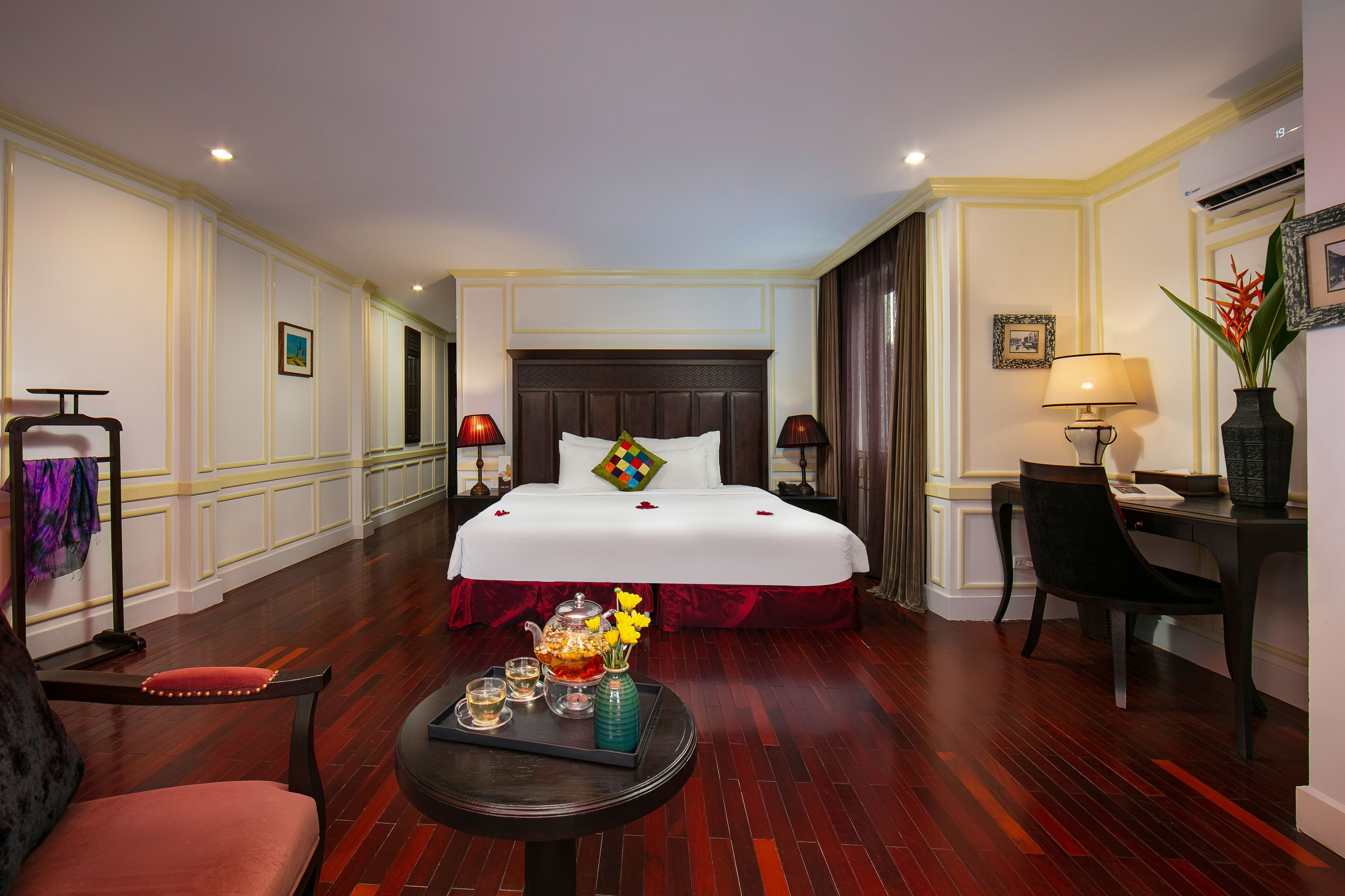 A hotel room with a large bed, a desk, a chair, a small table with a tray of drinks, and a vase with flowers.