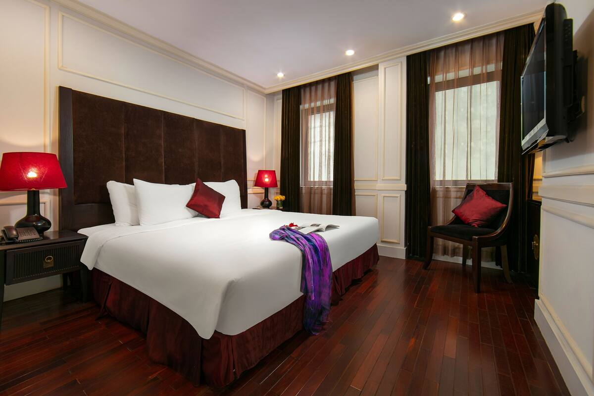 Habitación de hotel con una cama grande, dos lámparas de noche, un televisor montado en la pared y una silla con un cojín rojo.