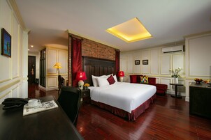 A hotel room with a large bed, a desk with a telephone and a magazine, a red chair, a small table with a vase of flowers, and a fireplace.