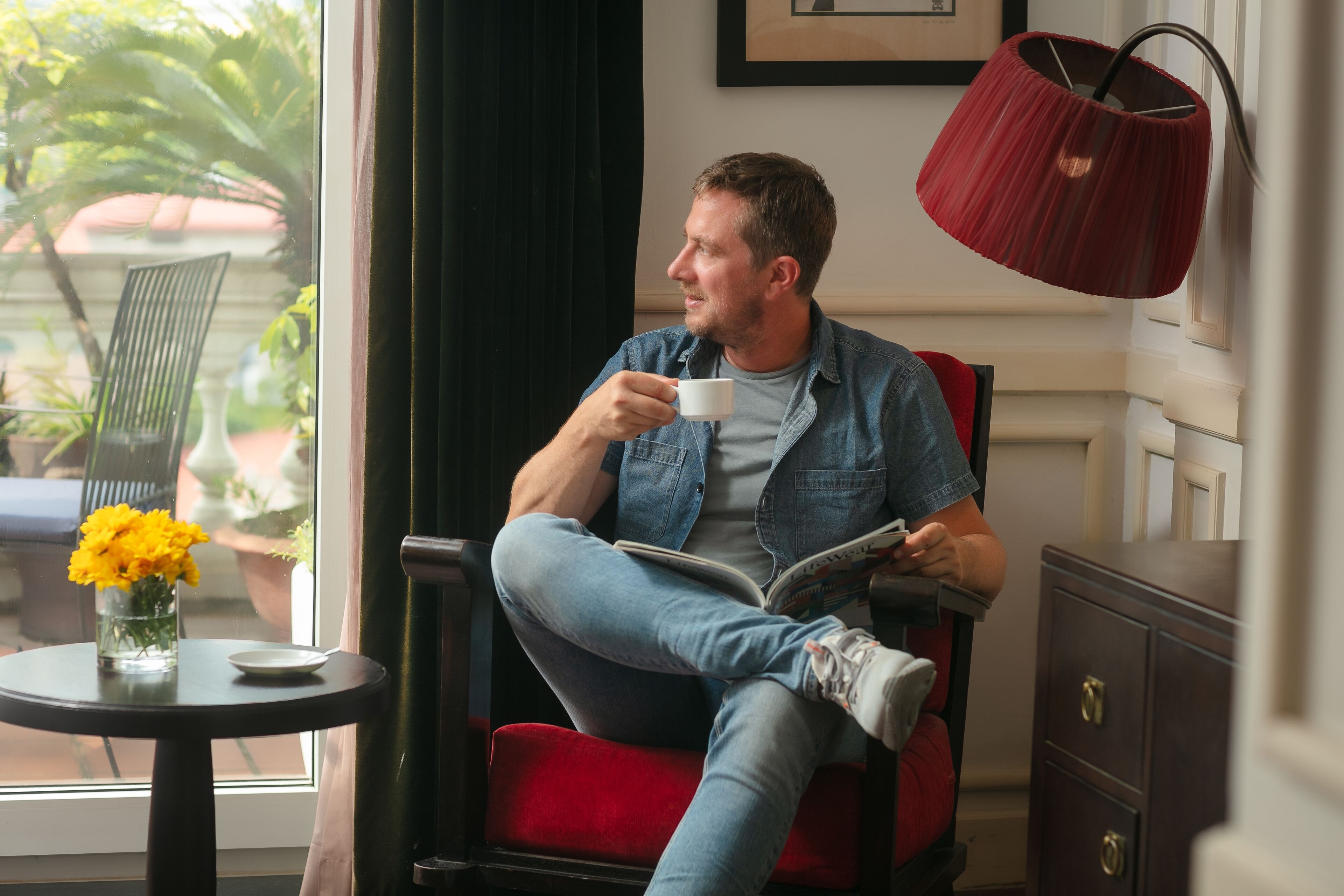 Ein Mann sitzt in einem roten Sessel, liest eine Zeitschrift und hält eine Kaffeetasse in der Hand. Neben ihm befindet sich ein Fenster mit Blick auf grüne Vegetation.