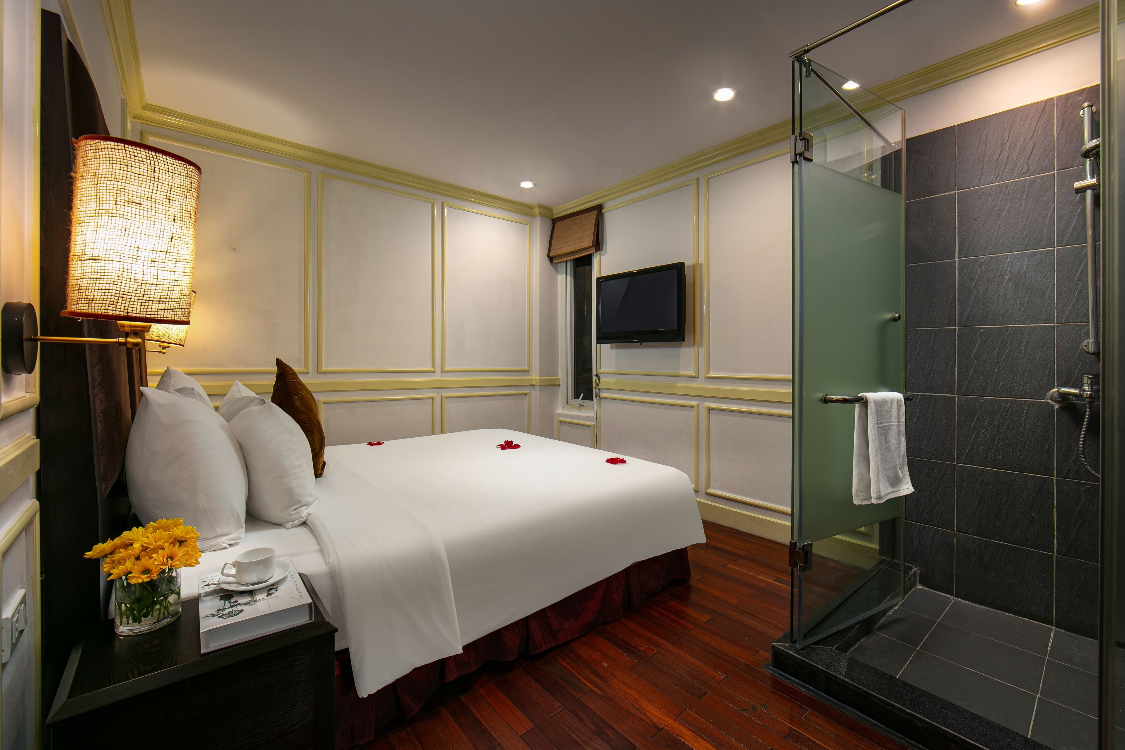 A hotel room with a bed, a bedside table with a vase of flowers, a television, and a glass-enclosed shower.