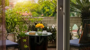 Una mesa redonda de vidrio con un libro, una taza blanca y un florero con flores amarillas en un balcón con sillas y plantas en macetas.