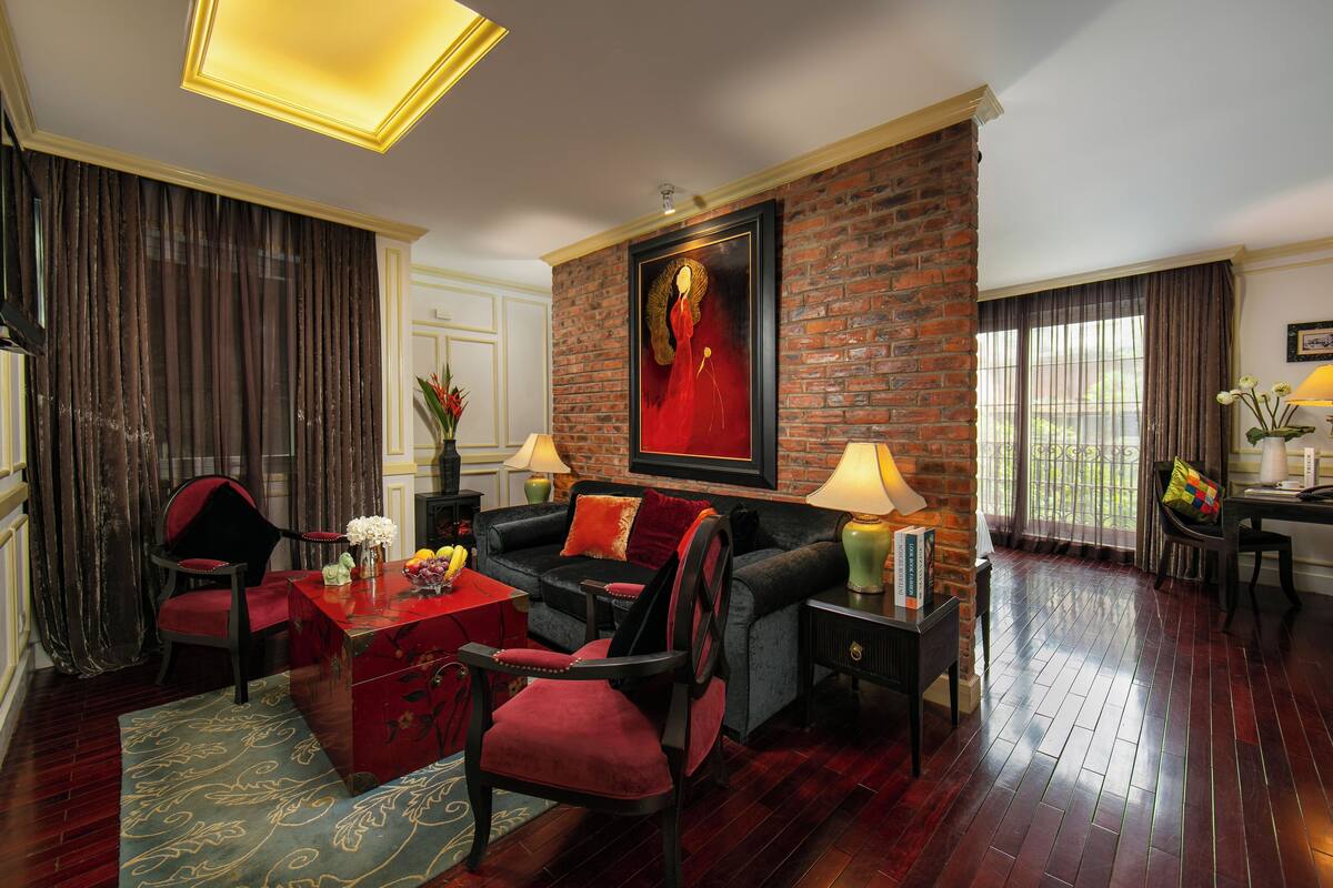 Una sala de estar con una pared de ladrillo, un sofá rojo, un sillón negro, una mesa de centro roja y una mesita auxiliar verde con una lámpara.