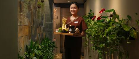 A corridor with stone walls, hanging plants, and a person holding a tray with a candle and incense sticks.