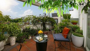 Una terraza en la azotea con macetas, una mesa de vidrio con un libro y flores, y una silla con cojín.