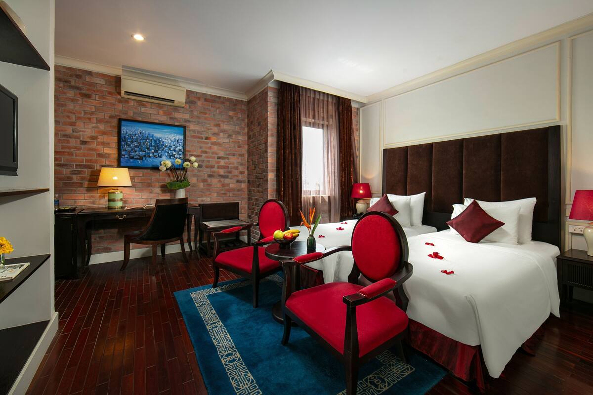Habitación de hotel con una cama grande, dos sillas rojas, una mesita con fruta, un escritorio y un televisor.