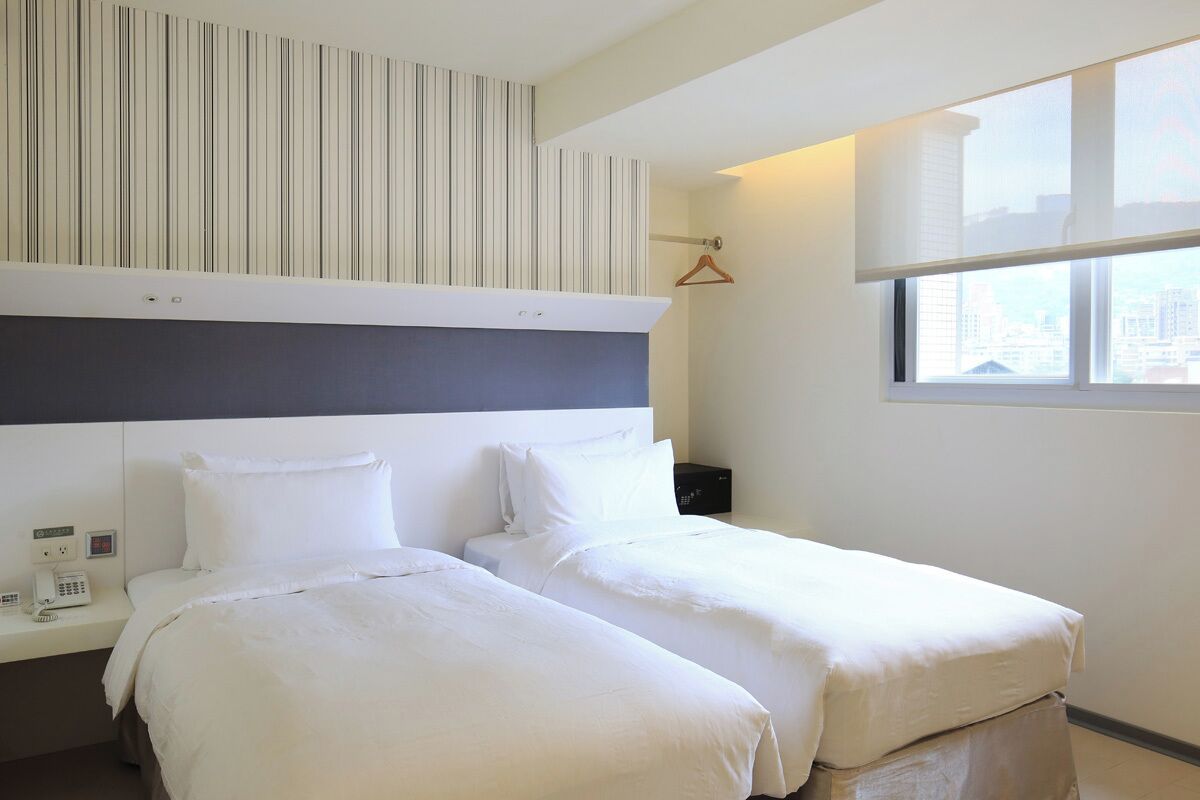 Une chambre d’hôtel avec deux lits, un mur à rayures, une fenêtre donnant sur la ville et une table de chevet avec un téléphone.