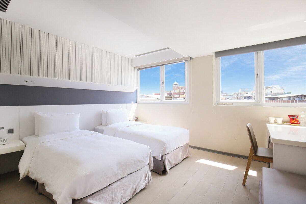 A hotel room with two beds, a desk, and a chair. There are large windows with a view of buildings.