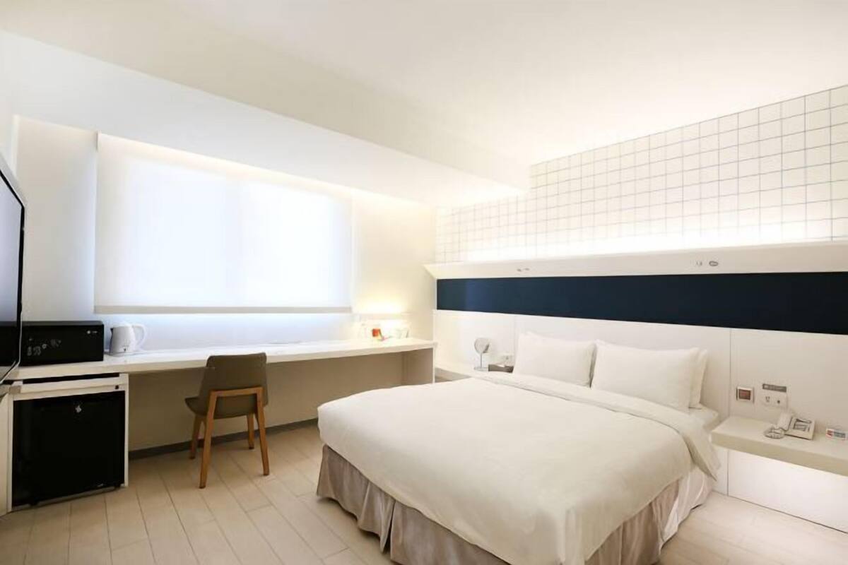 A hotel room with a bed, desk, chair, and a small kitchenette.