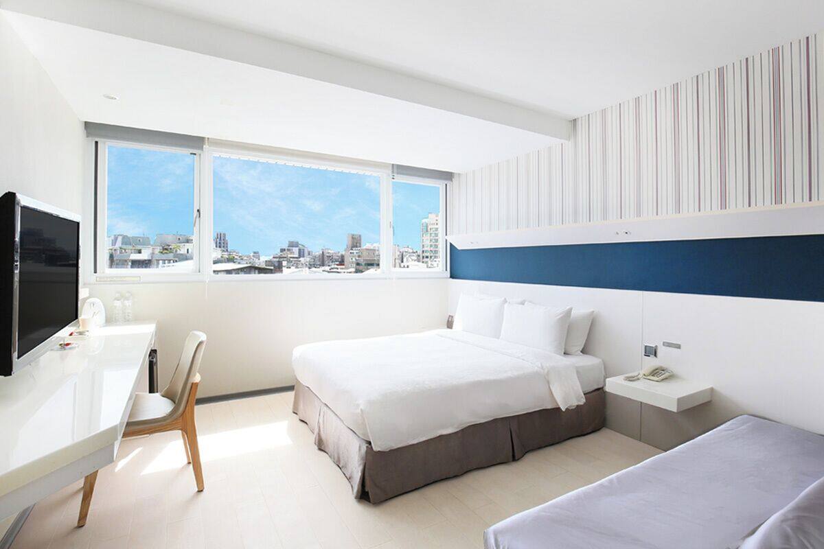A modern hotel room with a large bed, a desk, and a chair. There is a window with a city view.