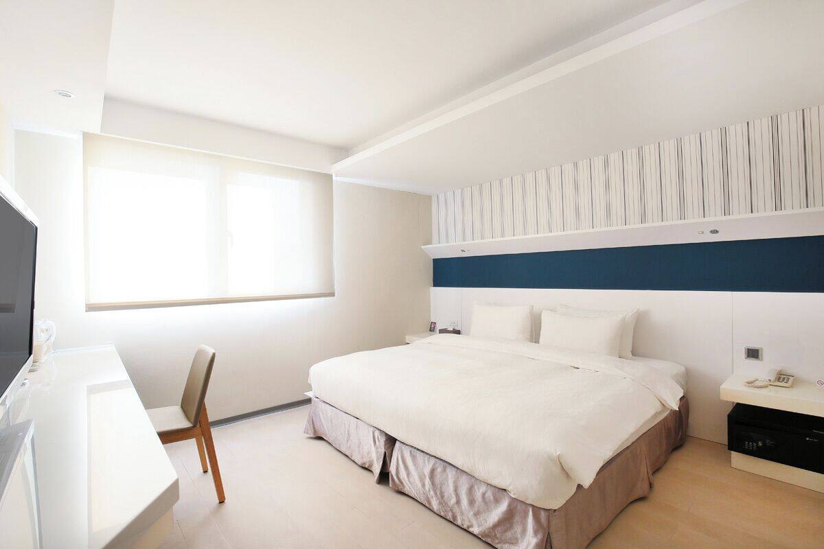 A modern hotel room with a large bed, a desk with a chair, a television, and a window providing natural light.