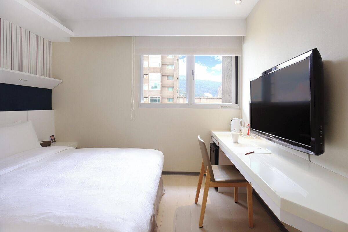 A hotel room with a bed, a desk with a flat-screen TV, a window, and a view of buildings.