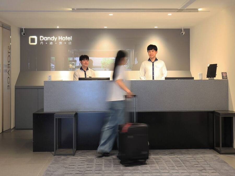 A reception desk at Dandy Hotel with two staff members, a guest with luggage, and a modern interior design.
