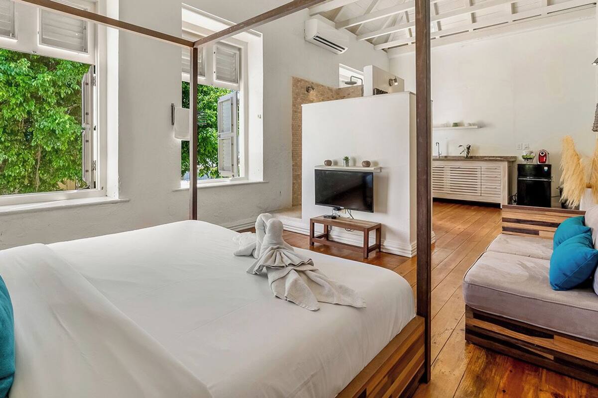 A bedroom with a large bed, a TV, a wooden floor, and a view of greenery outside.