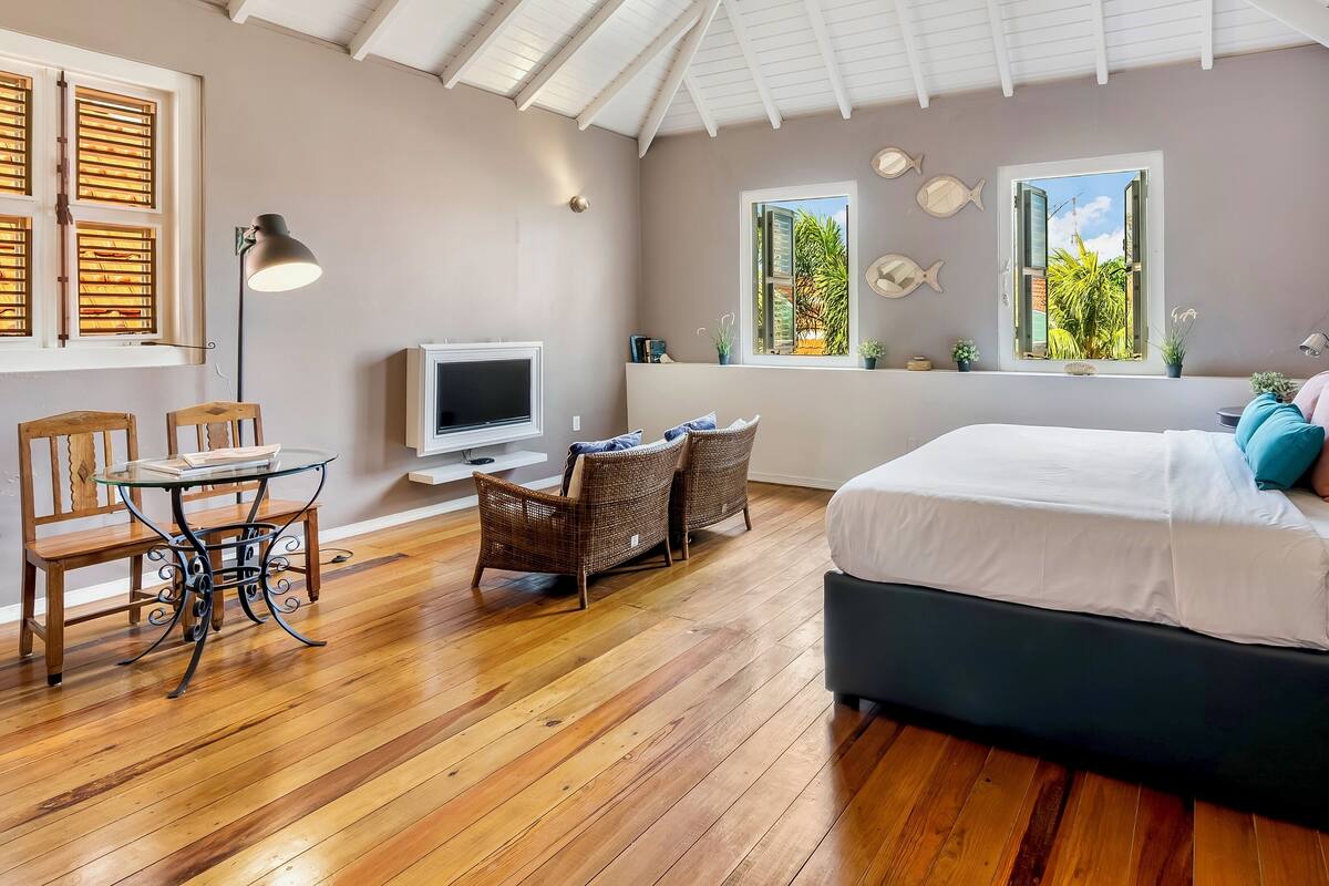 A bedroom with a bed, a TV, a dining area with chairs, and windows with views of greenery.