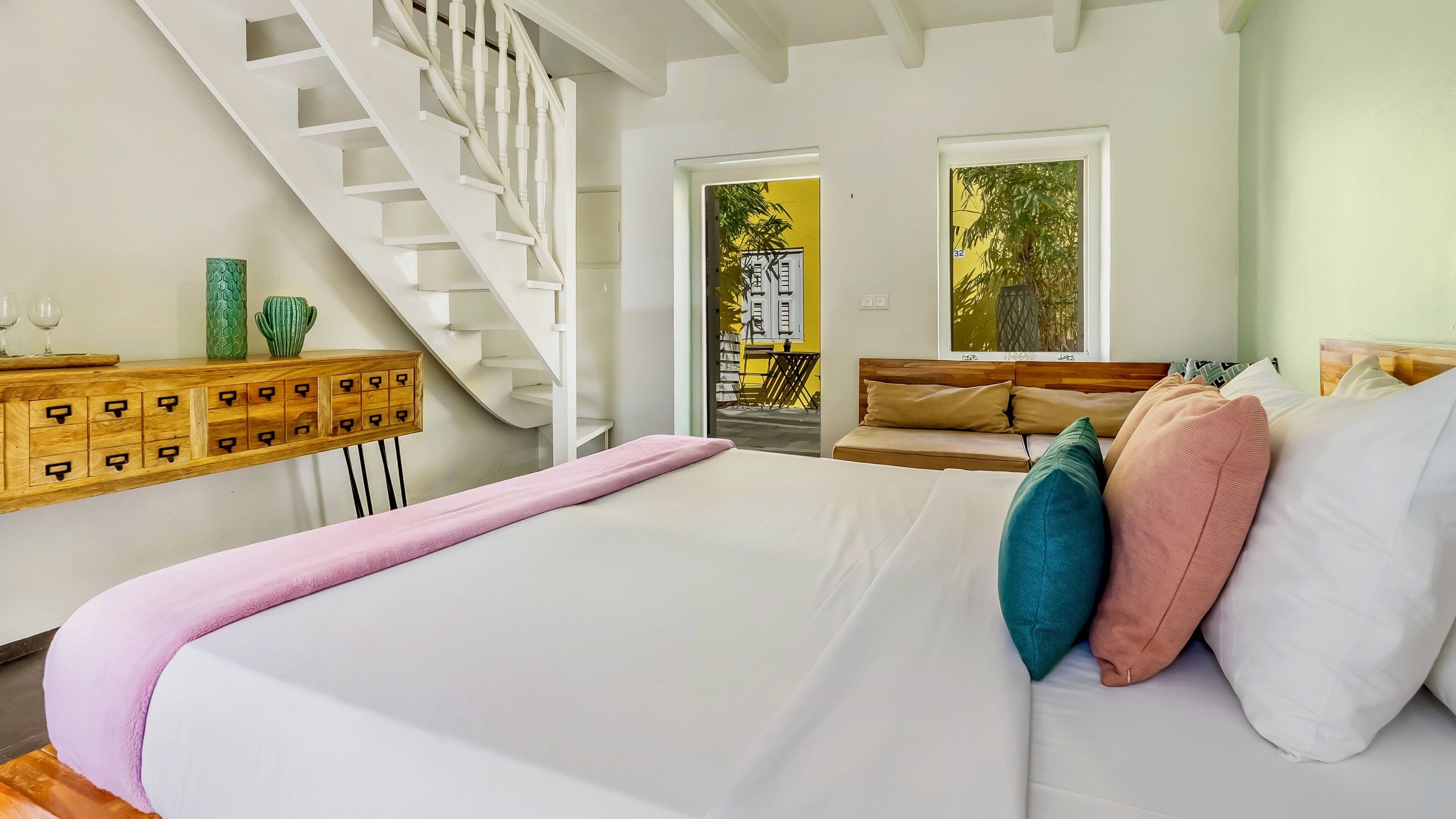 Un dormitorio con una cama grande, un aparador de madera con cajones, una escalera blanca y vistas al exterior por una ventana.