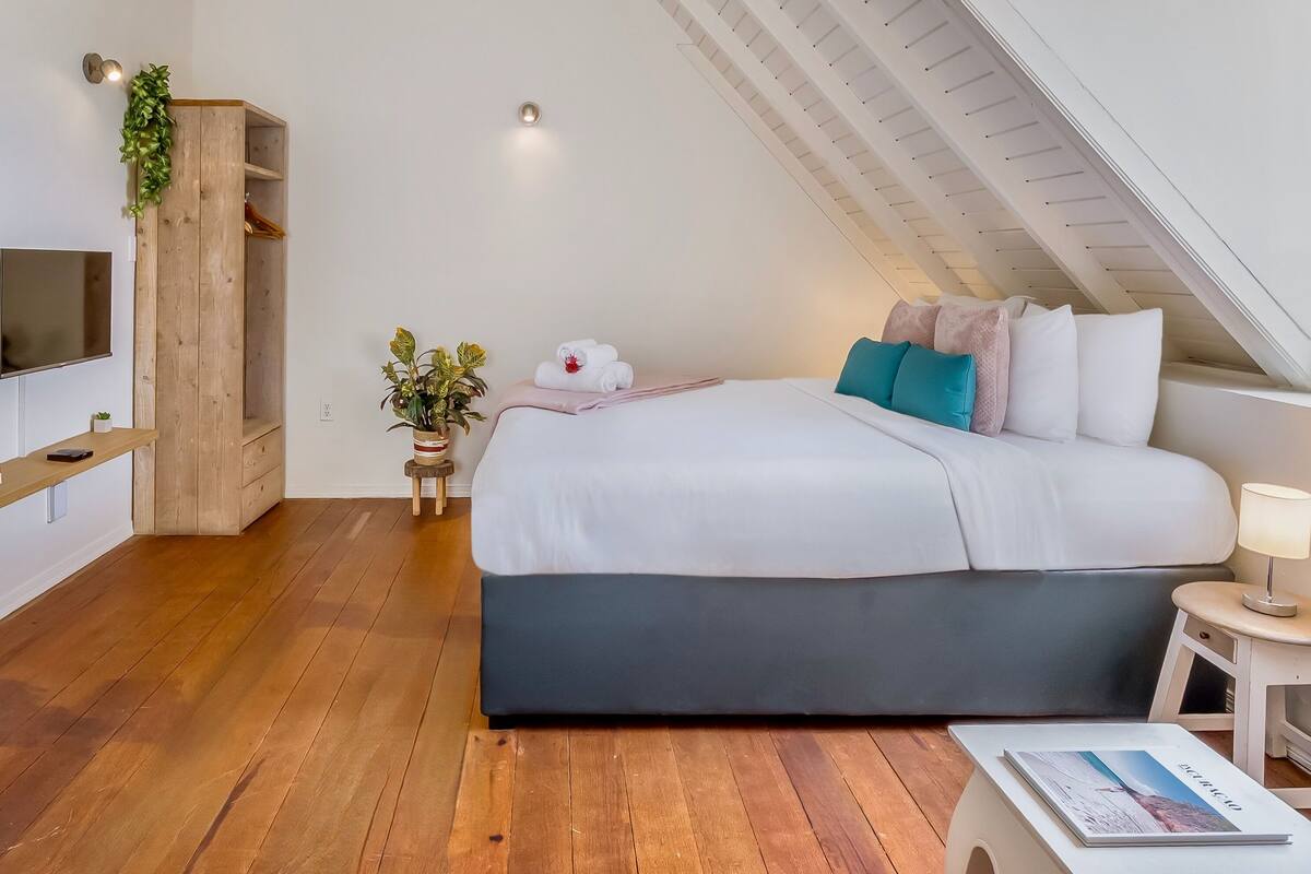 A modern bedroom with a large bed, a TV mounted on the wall, a wooden shelf, a small plant, and a bedside table with a lamp.