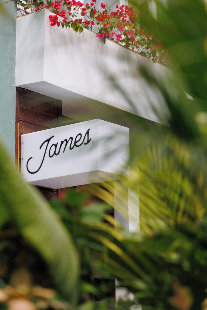 Un letrero blanco con el nombre 'James' en cursiva, colgado de un balcón con flores rojas.