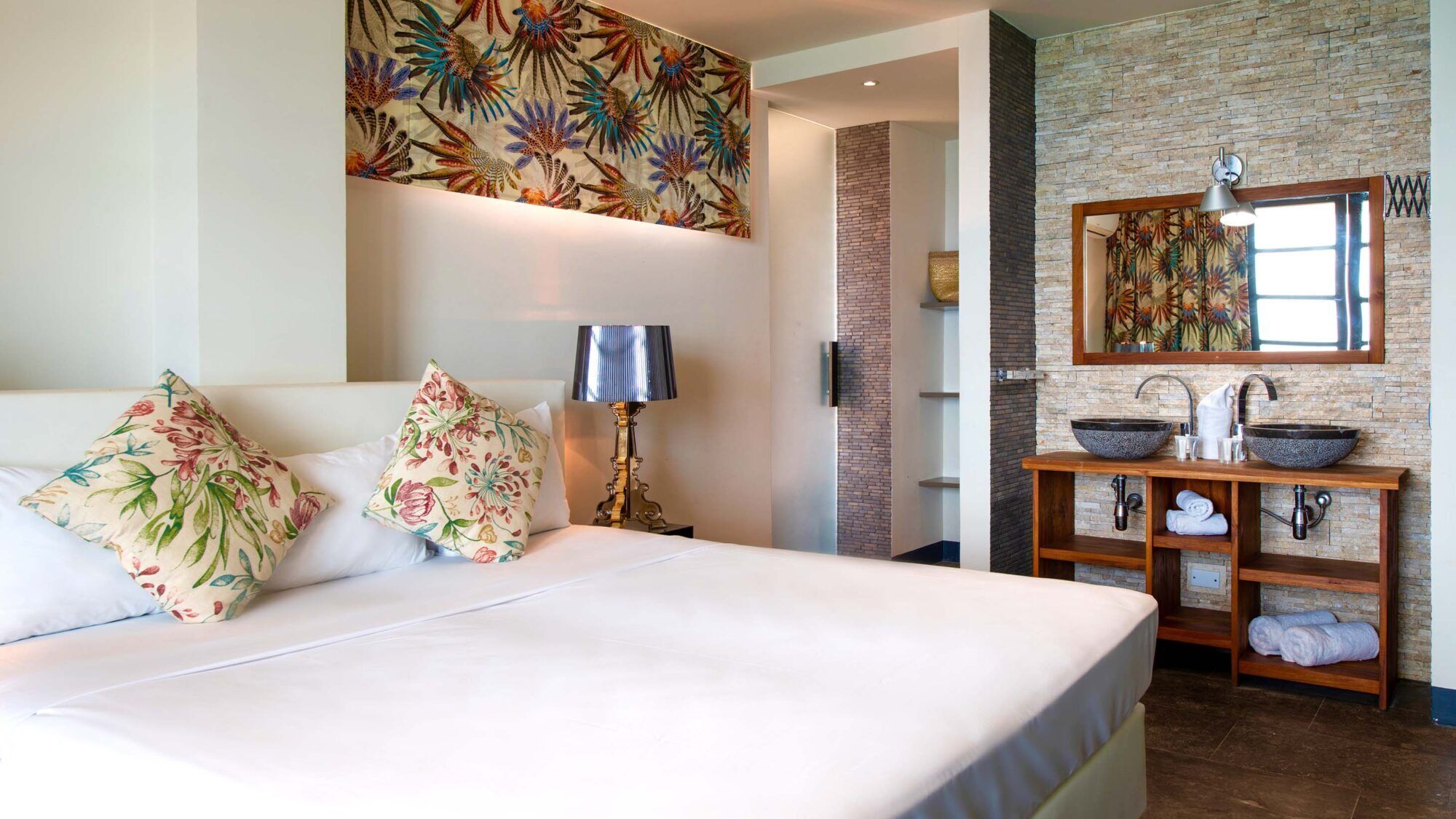 Een hotelkamer met een groot bed, een nachtkastje met lamp, een badkamer met een stenen wastafel en een spiegel.