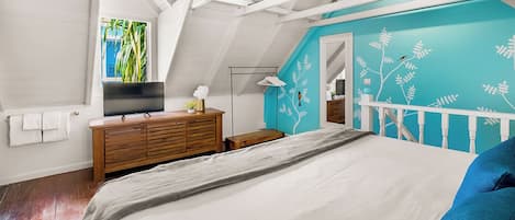 Une chambre à coucher comprenant un lit, une commode en bois, un téléviseur et un mur d’accent de couleur turquoise orné d’un motif d’arbre.