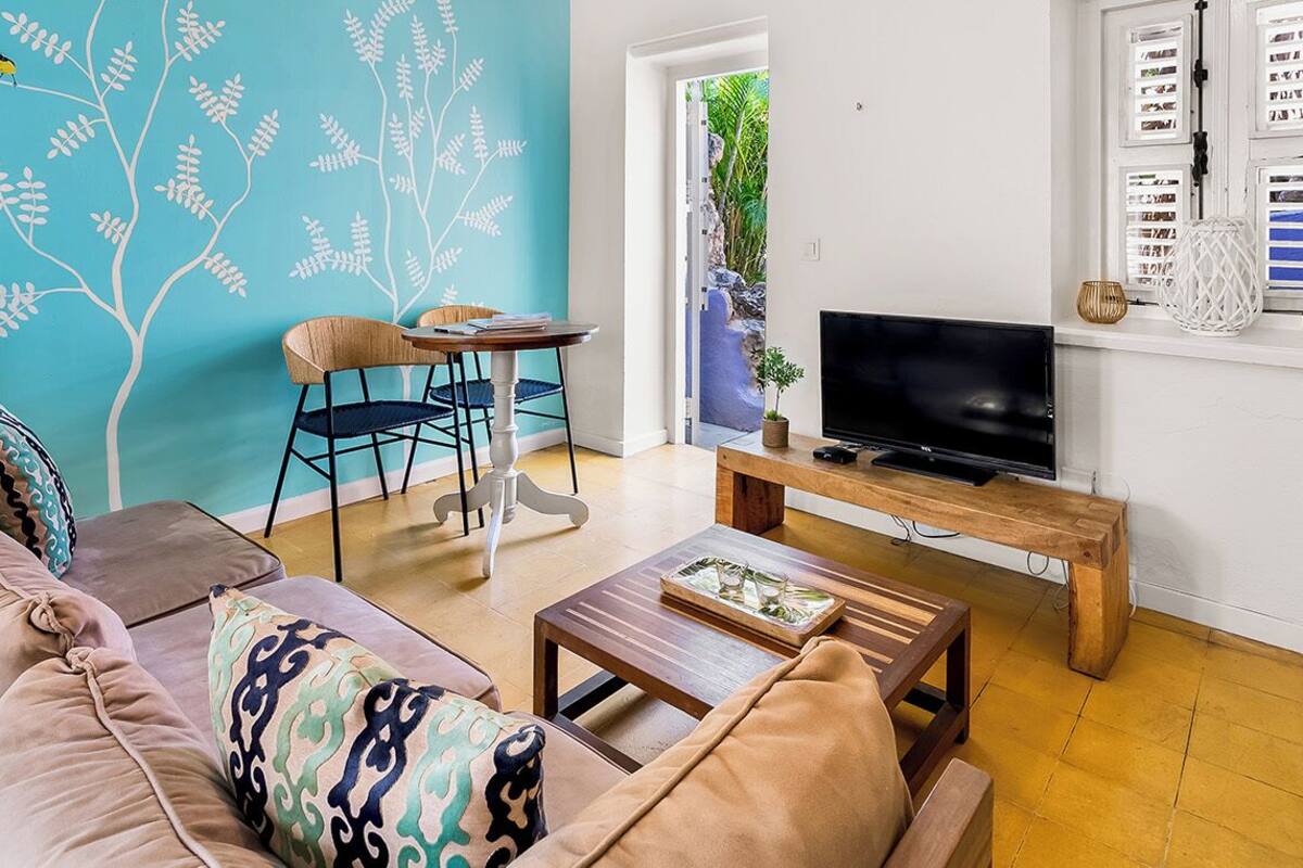 A living room with a sofa, chairs, a coffee table, a TV on a wooden stand, and a wall with a tree mural.