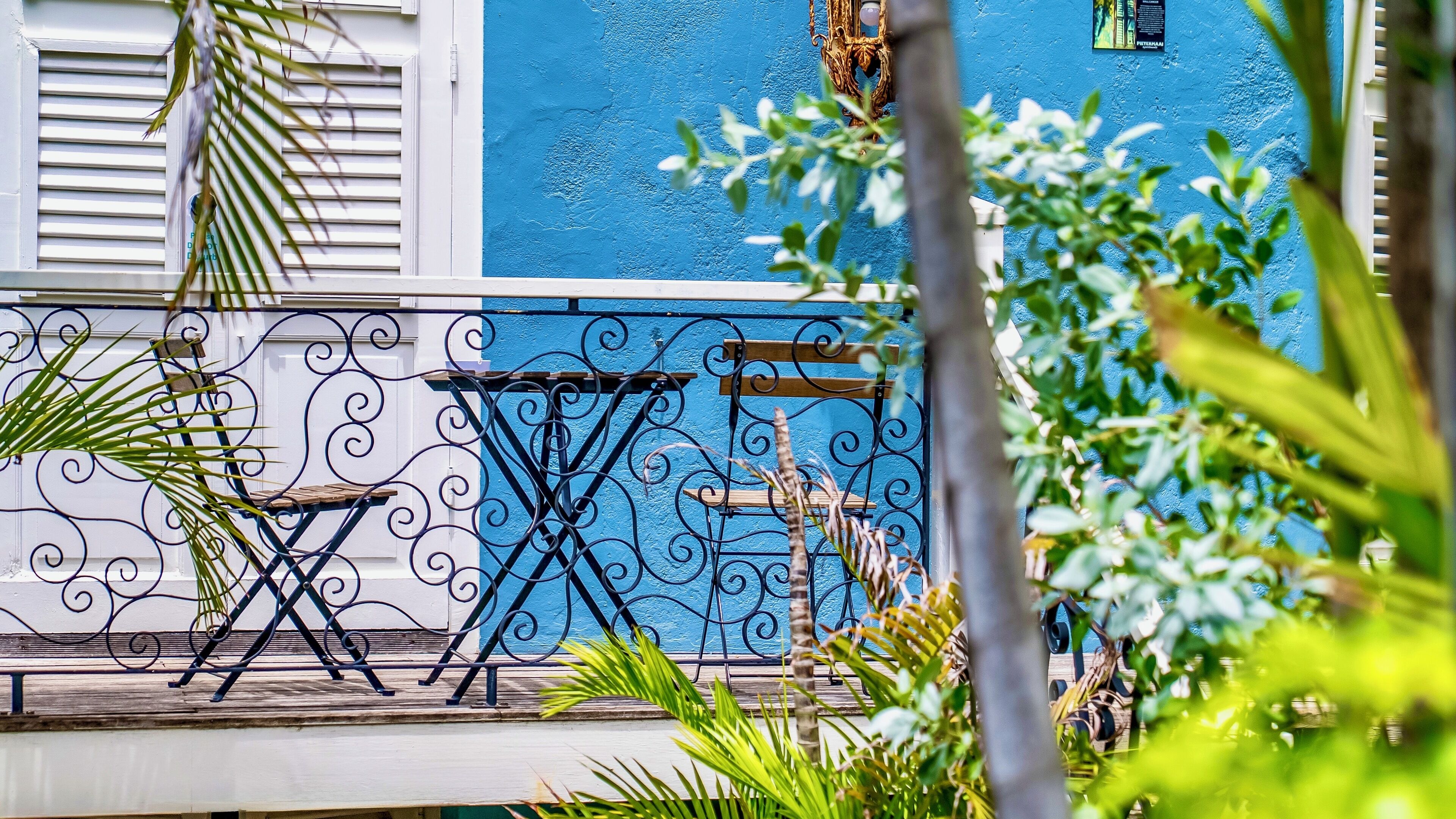 Un balcón con pared azul, baranda de metal y una silla de madera.