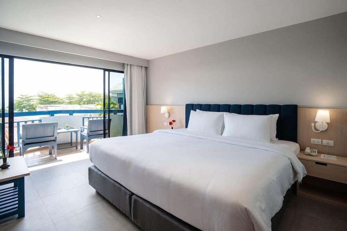 A hotel room with a large bed, a balcony with chairs, and a view of greenery.