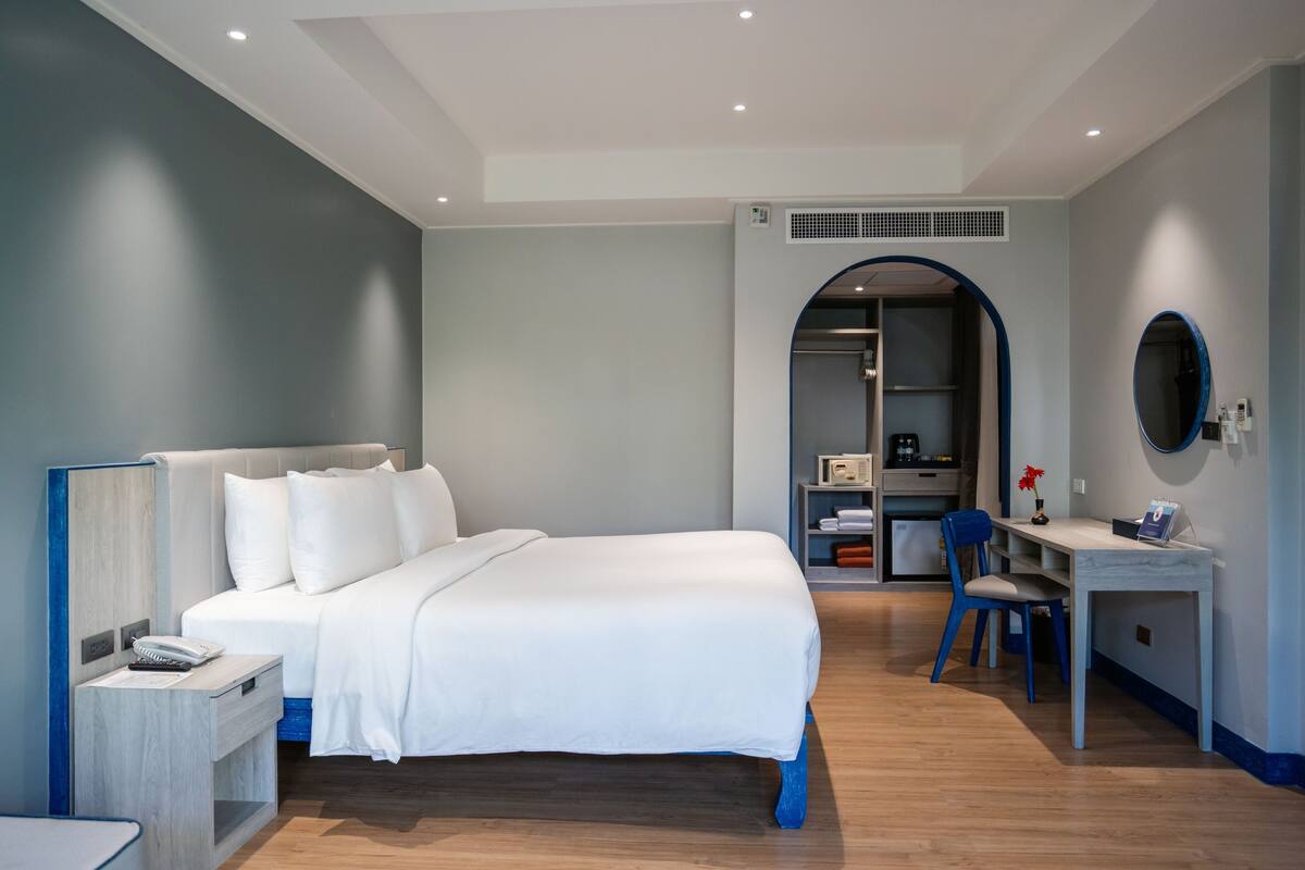 A modern hotel room with a large bed, a desk, and a chair. There is a wardrobe and a small seating area with a table and chairs.