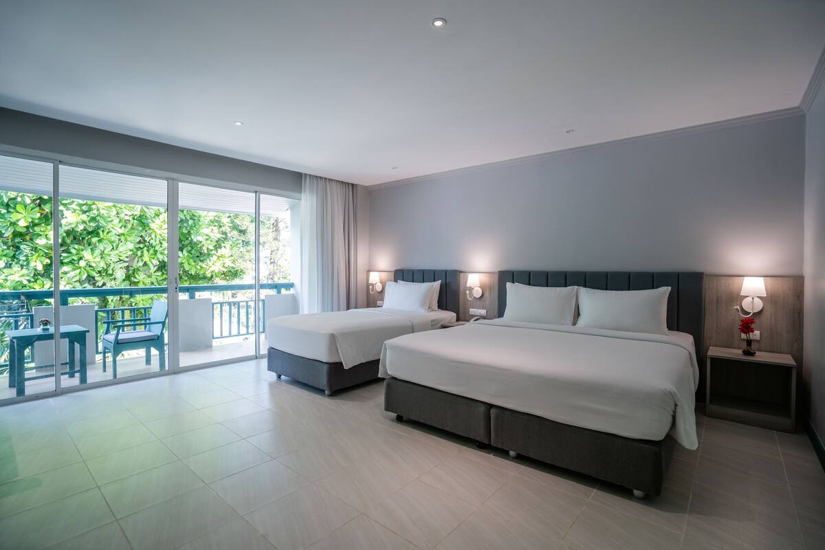A modern hotel room with two beds, a balcony with a table and chairs, and a view of greenery.
