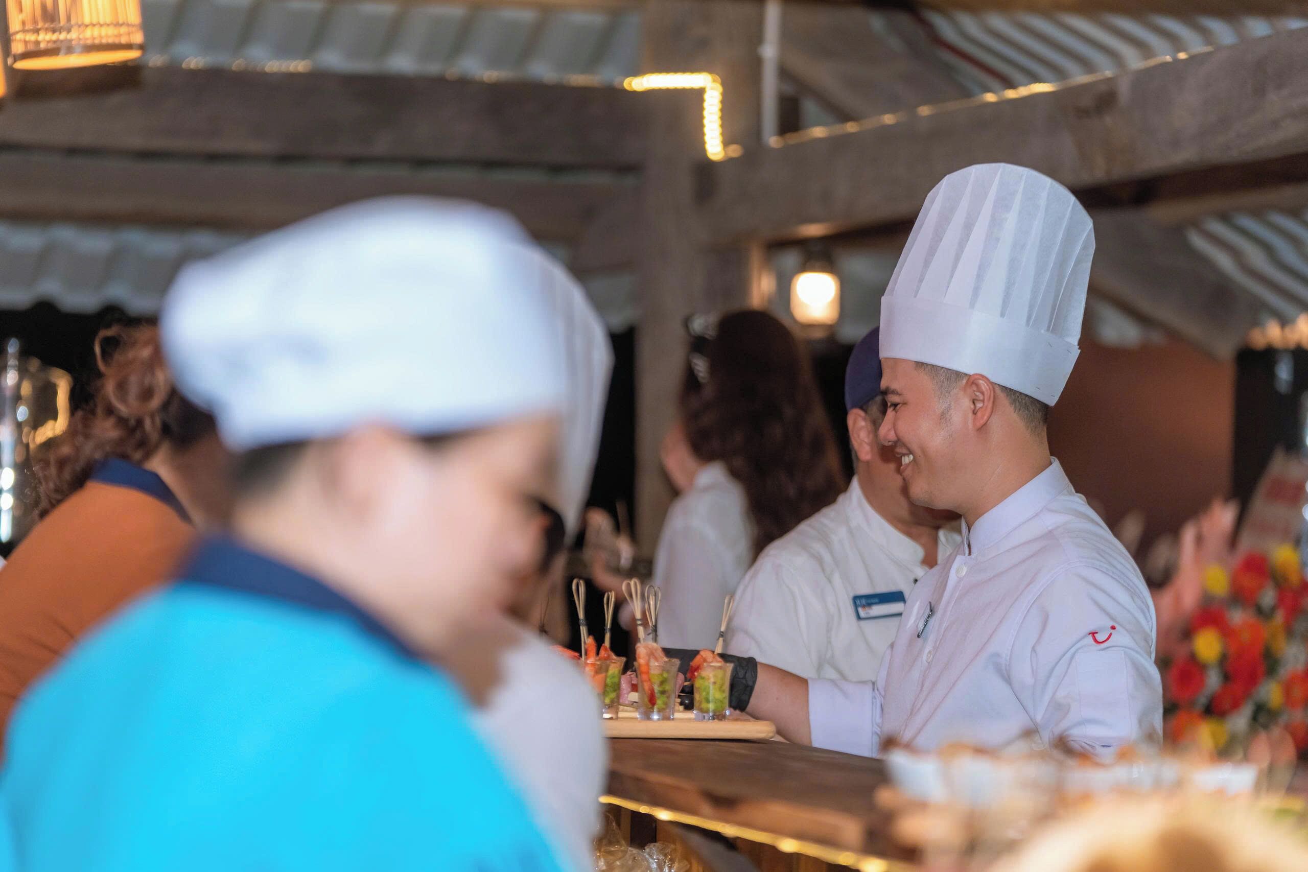 A chef in a white uniform with a chef's hat, standing at a counter with a tray of food items.