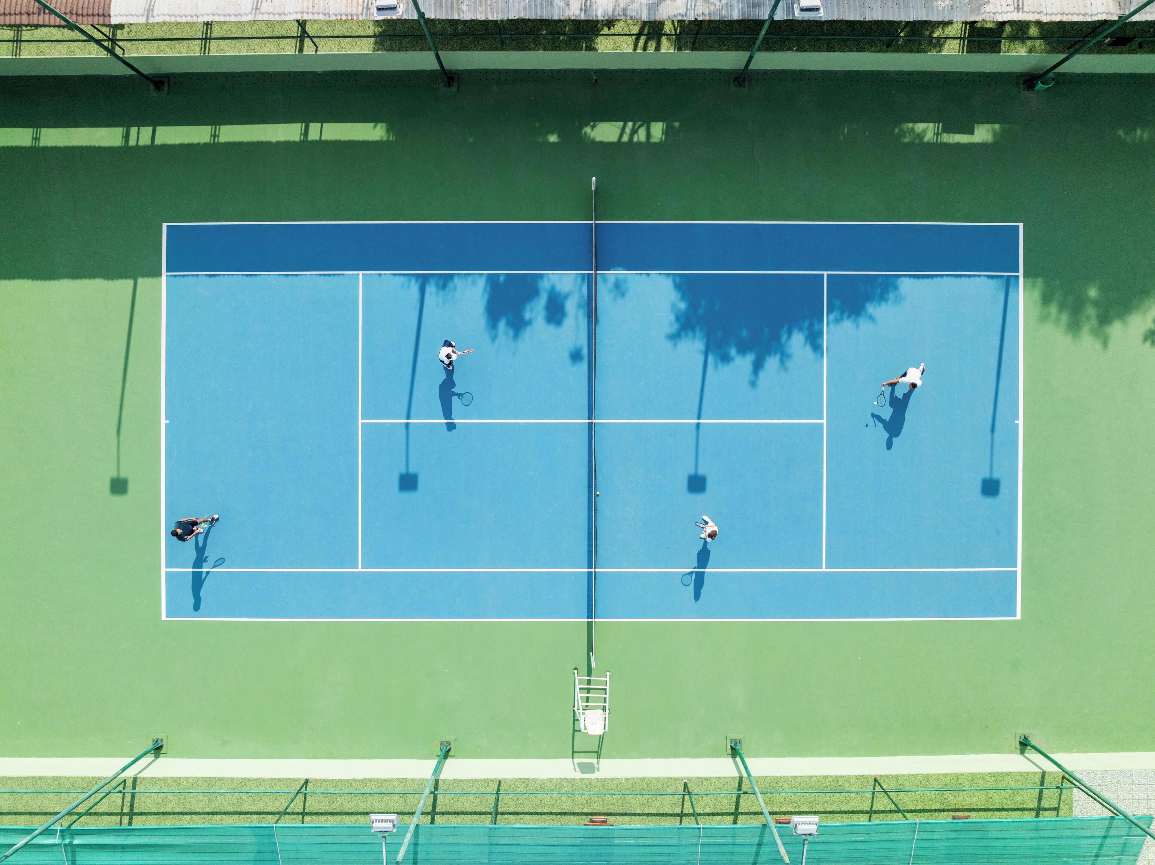 An overhead view of a tennis court with four players, green and blue court surfaces, and surrounding green fencing.