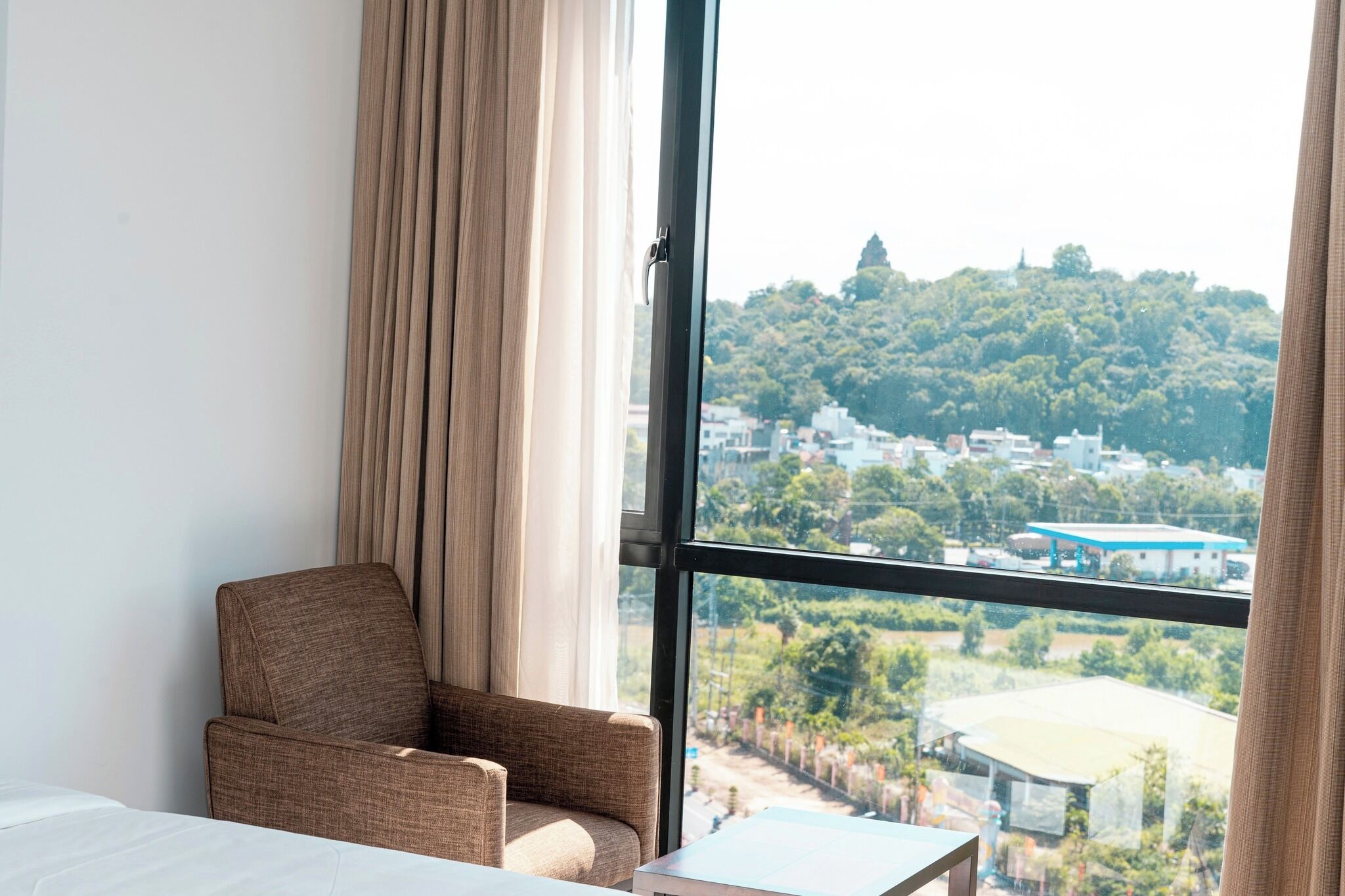 A hotel room with a large window, a brown armchair, a small table, and a bed.