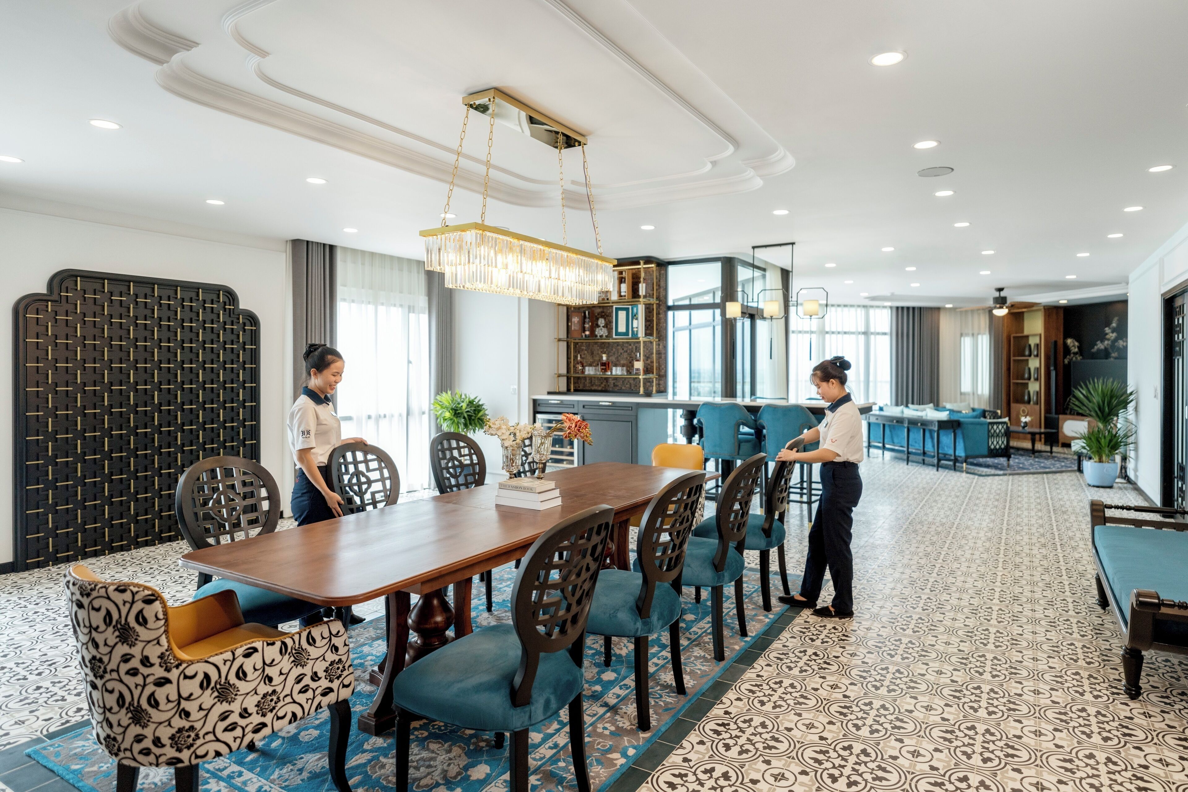 A hotel dining area with a long wooden table, blue upholstered chairs, and a chandelier.