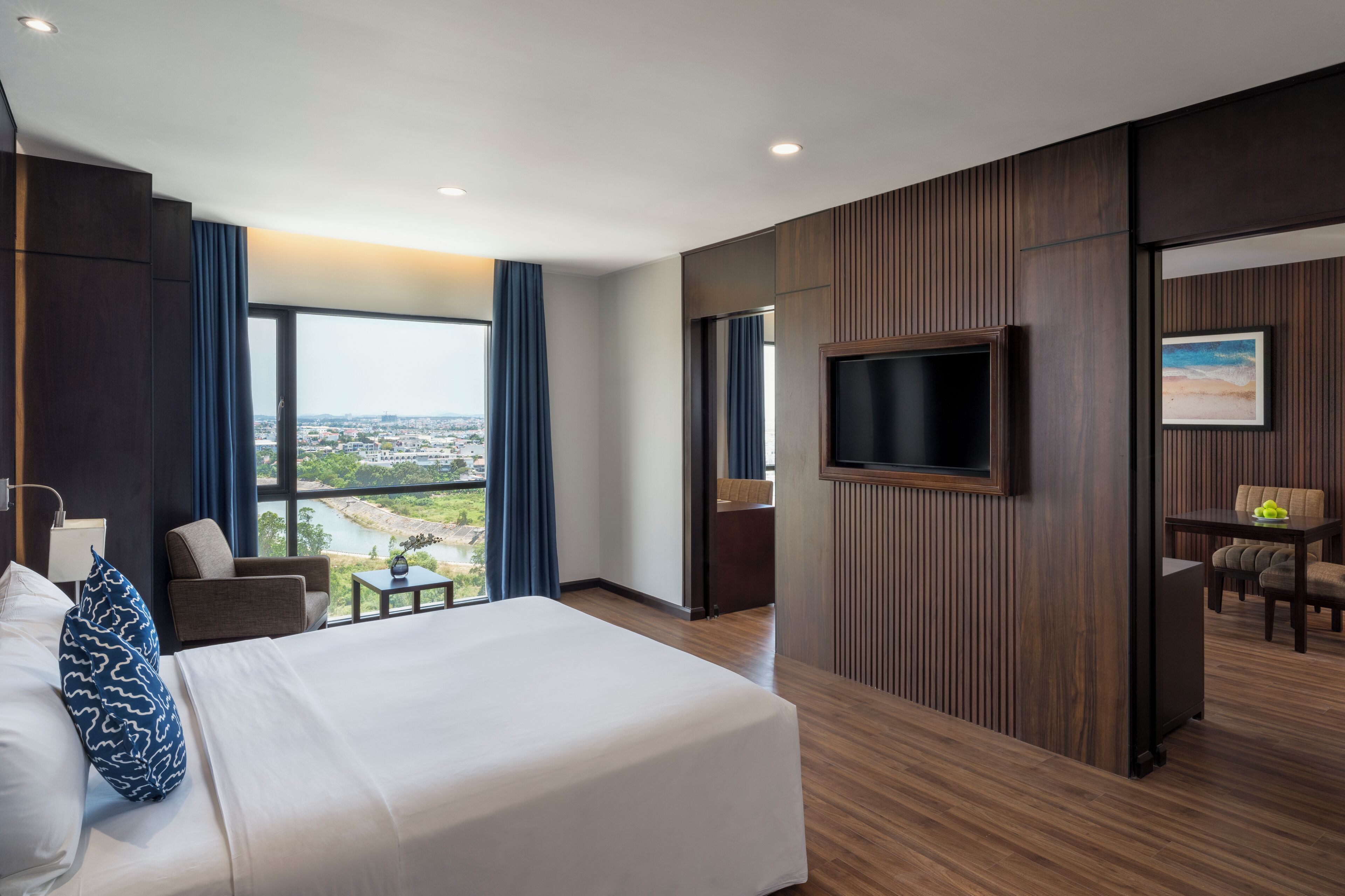 A modern hotel room with a large bed, a TV, a dining area, and a view of a cityscape.