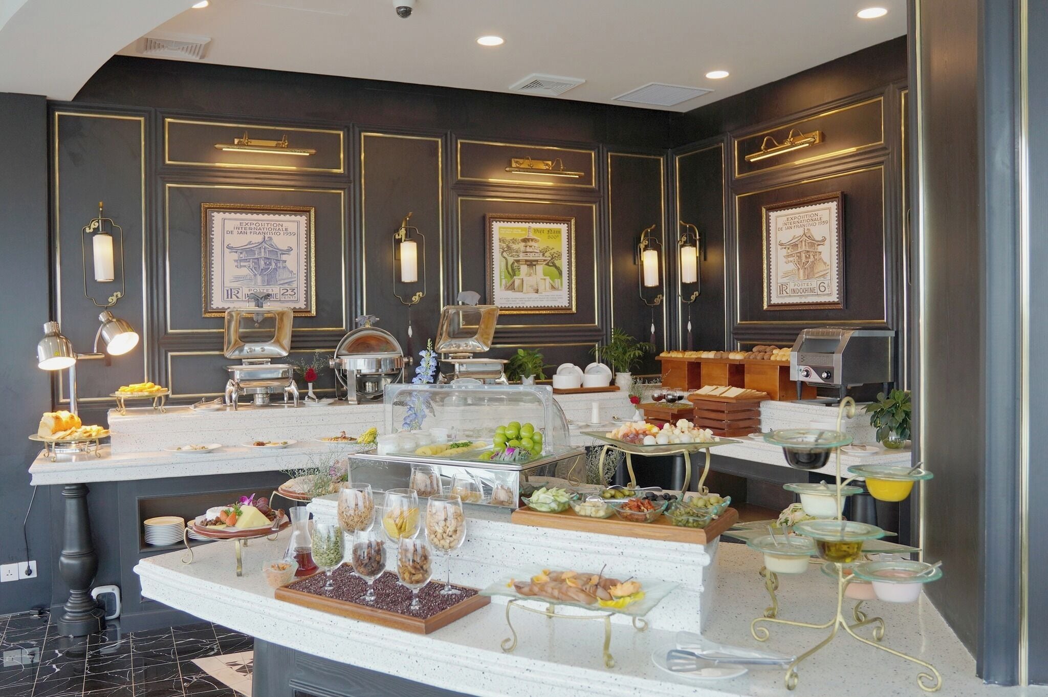 A buffet setup with various food items and beverages, framed pictures on the wall, and a marble floor.