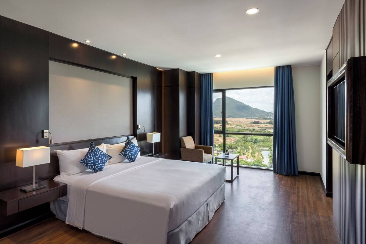 Een moderne hotelkamer met een groot bed, nachtkastjes, een stoel en uitzicht op een bergachtig landschap door grote ramen.