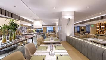 A modern restaurant with long communal tables, cushioned seating, and a view of the cityscape.