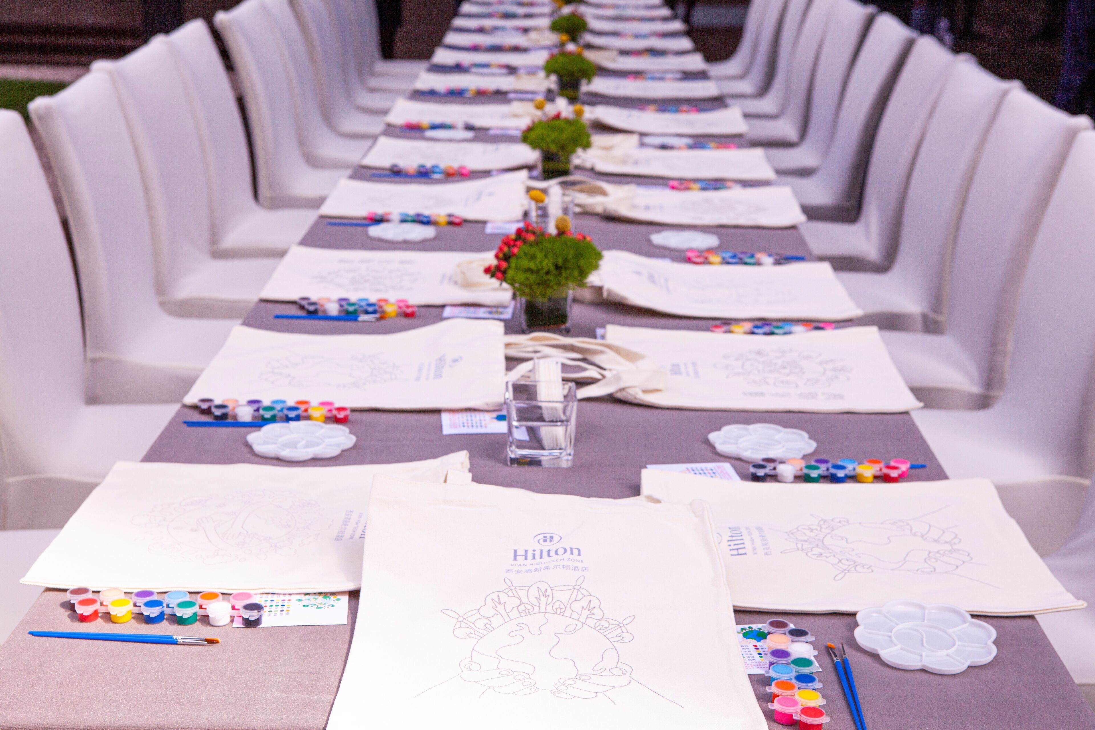 Een lange tafel klaargezet met kunstbenodigdheden en lege vellen papier, waarschijnlijk voor een schildersactiviteit.