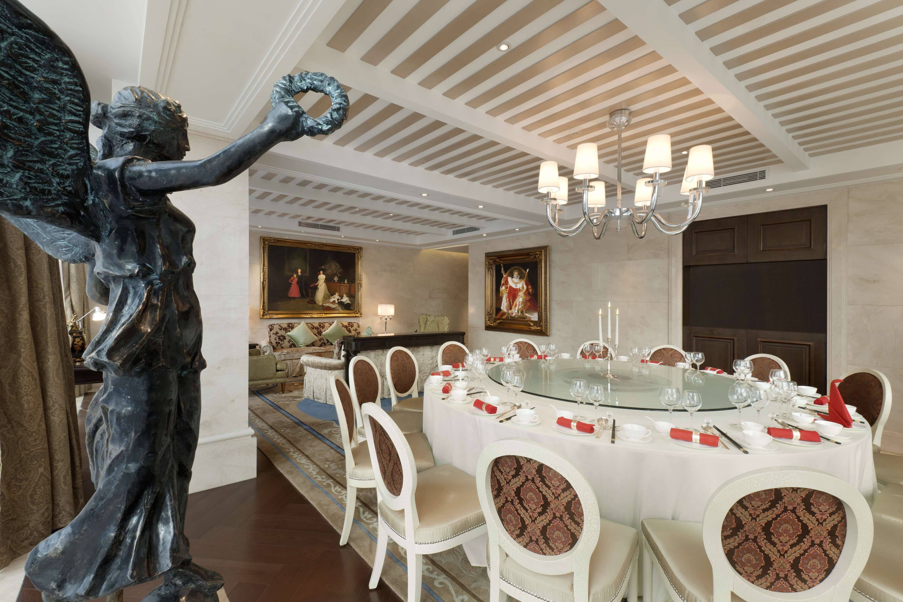 Un comedor formal con una mesa redonda de cristal dispuesta para una comida, rodeada de sillas blancas con tapicería estampada. Una estatua de bronce de una figura sosteniendo una corona se encuentra a la izquierda. La habitación presenta un techo alto con un diseño a rayas y un candelabro.