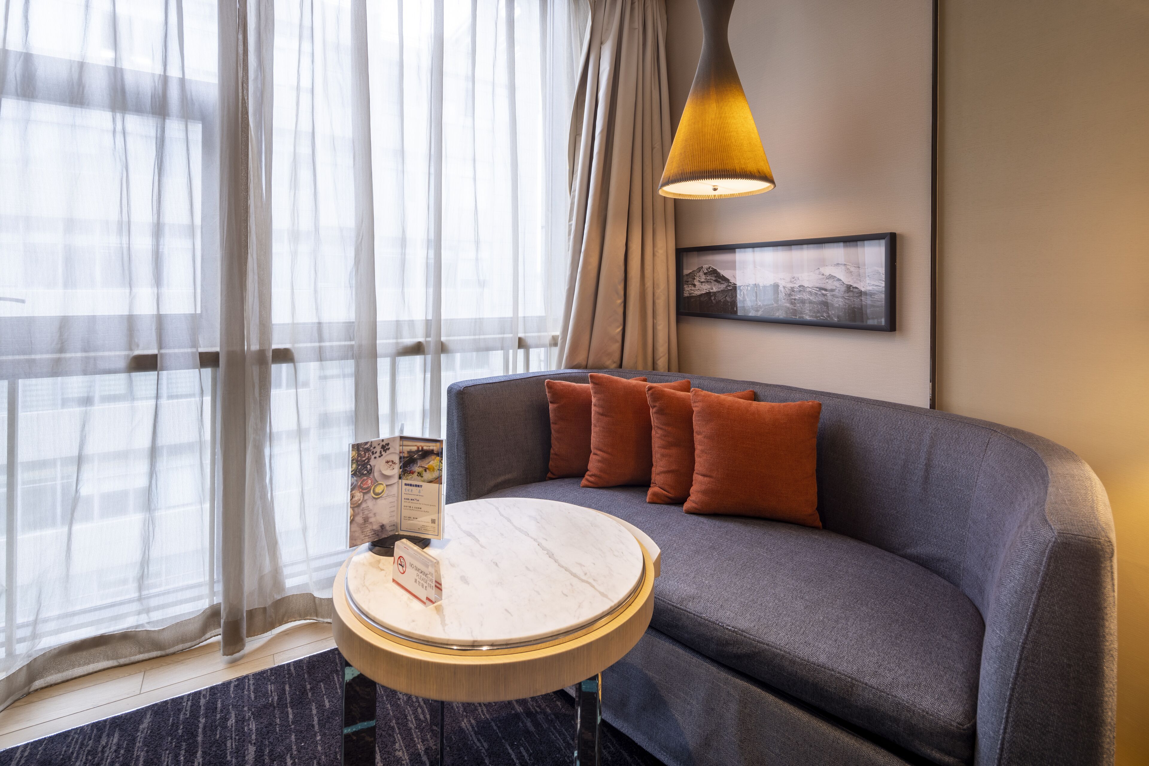 A modern hotel room with a grey sofa, a round wooden side table, sheer curtains, a framed picture on the wall, and a hanging pendant light.