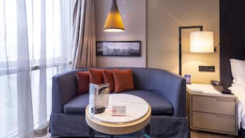 A hotel room with a curved grey sofa, a small round table with a magazine, a bed with white linens, a nightstand with a lamp, and a framed picture on the wall.