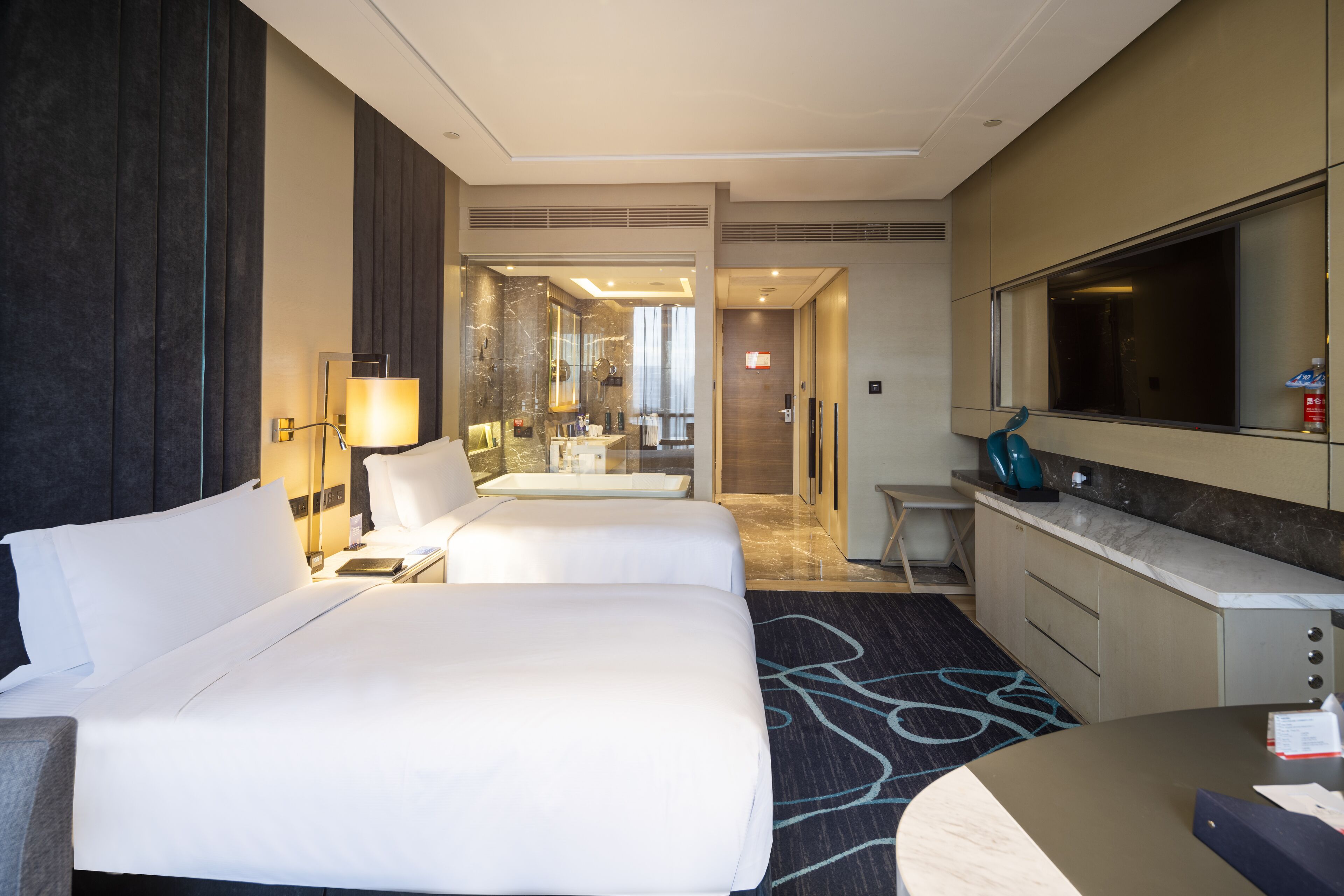 A hotel room with two beds, a desk, a mirror, and a bathroom visible through an open door.
