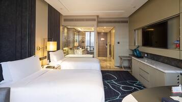 Ein Hotelzimmer mit zwei Betten, einem Schreibtisch, einem Spiegel und einem durchgehenden Türbogen einsehbarer Badezimmer.