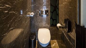 A modern bathroom with a marble floor, a white toilet, a trash bin, and a towel dispenser.