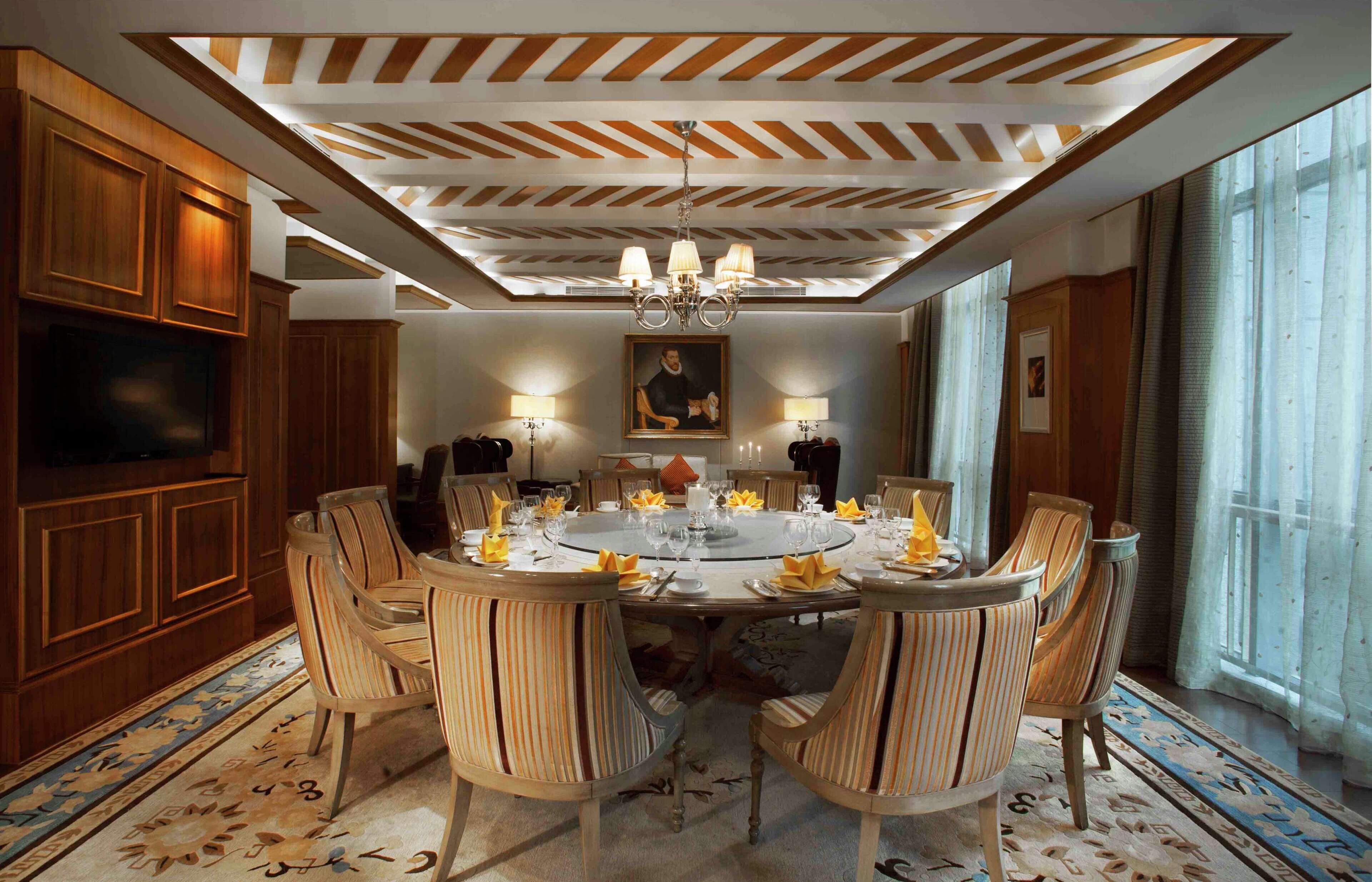 A dining room with a round table, six chairs, and a chandelier. There is a television on the left and a painting on the wall.