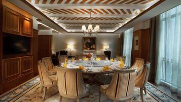 A dining room with a round table, six chairs, and a chandelier. There is a television on the left and a painting on the wall.