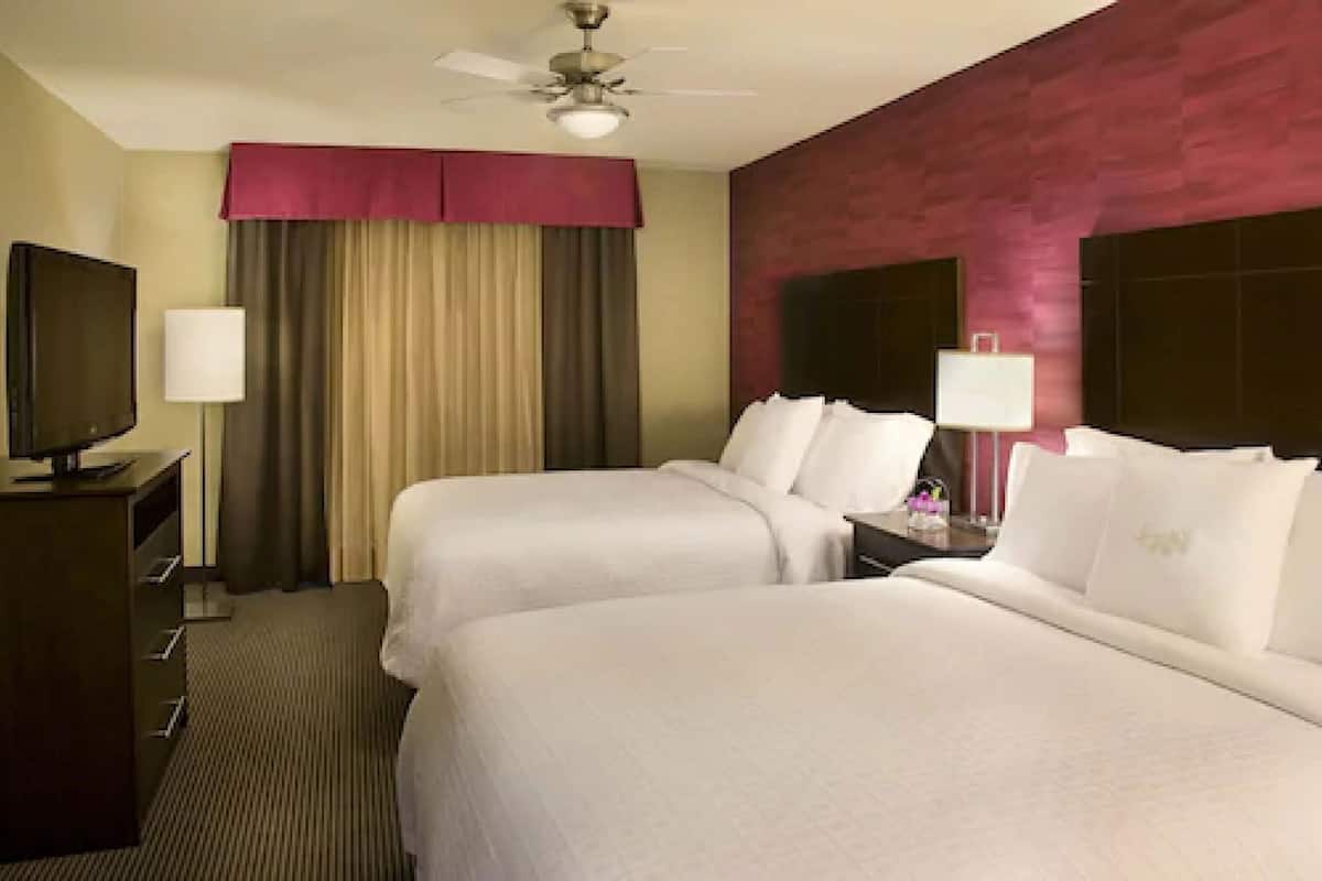 A hotel room with two beds, a television, a desk with a lamp, and a ceiling fan.