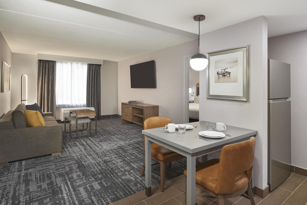 A hotel room with a gray sofa, a dining table with chairs, a small kitchenette, and a large window with curtains.
