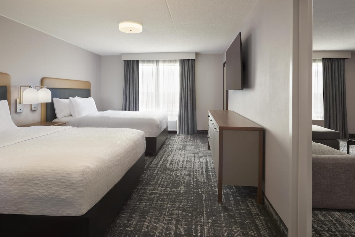 A hotel room with two beds, a desk, a flat-screen TV, and a large window with curtains.