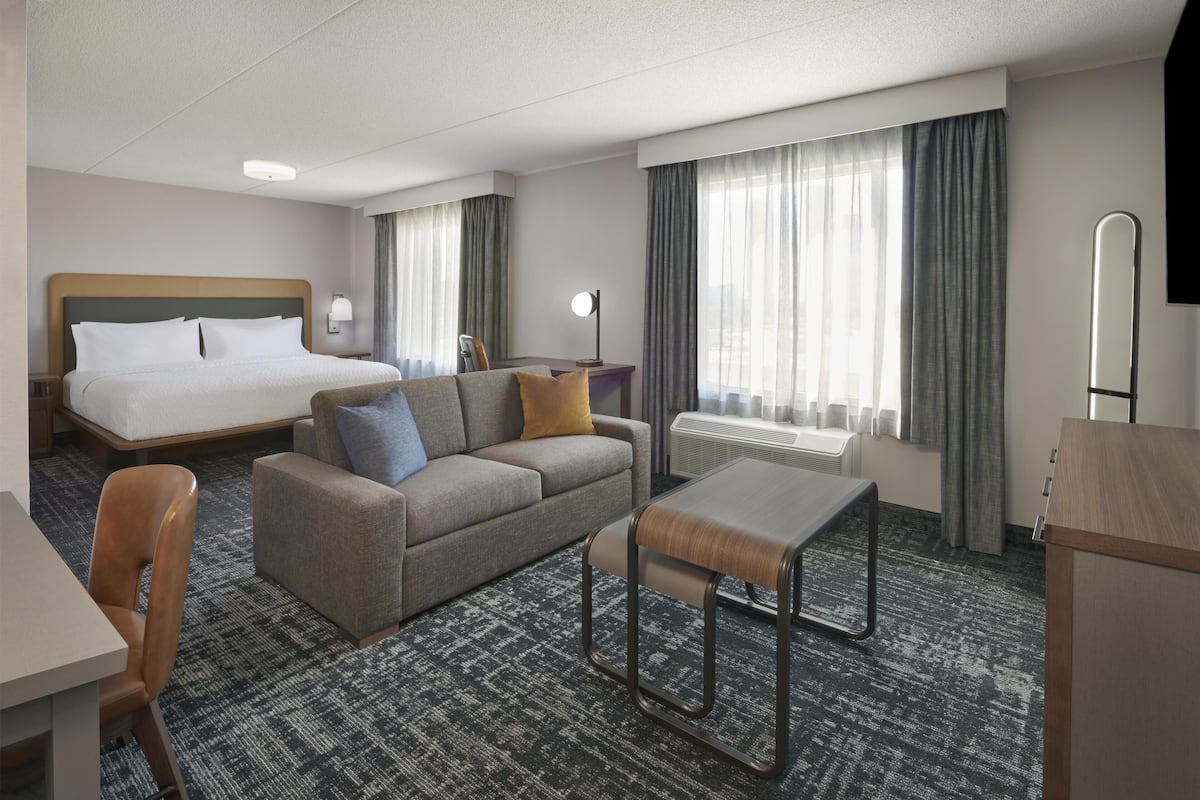 A hotel room with a bed, a sofa, a table, a chair, a desk, a lamp, a mirror, and a window with curtains.